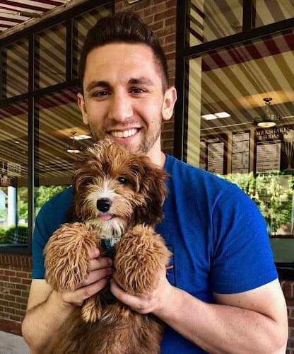 Guy grins as he holds a little puppy in his arms. He stands outside of a brick building and wears a royal blue t-shirt.