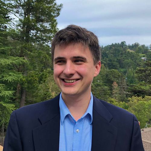 Jack smiles and looks off-camera. He wears a blue button-up and a navy jacket. A forest and blue sky fill the background behind him.